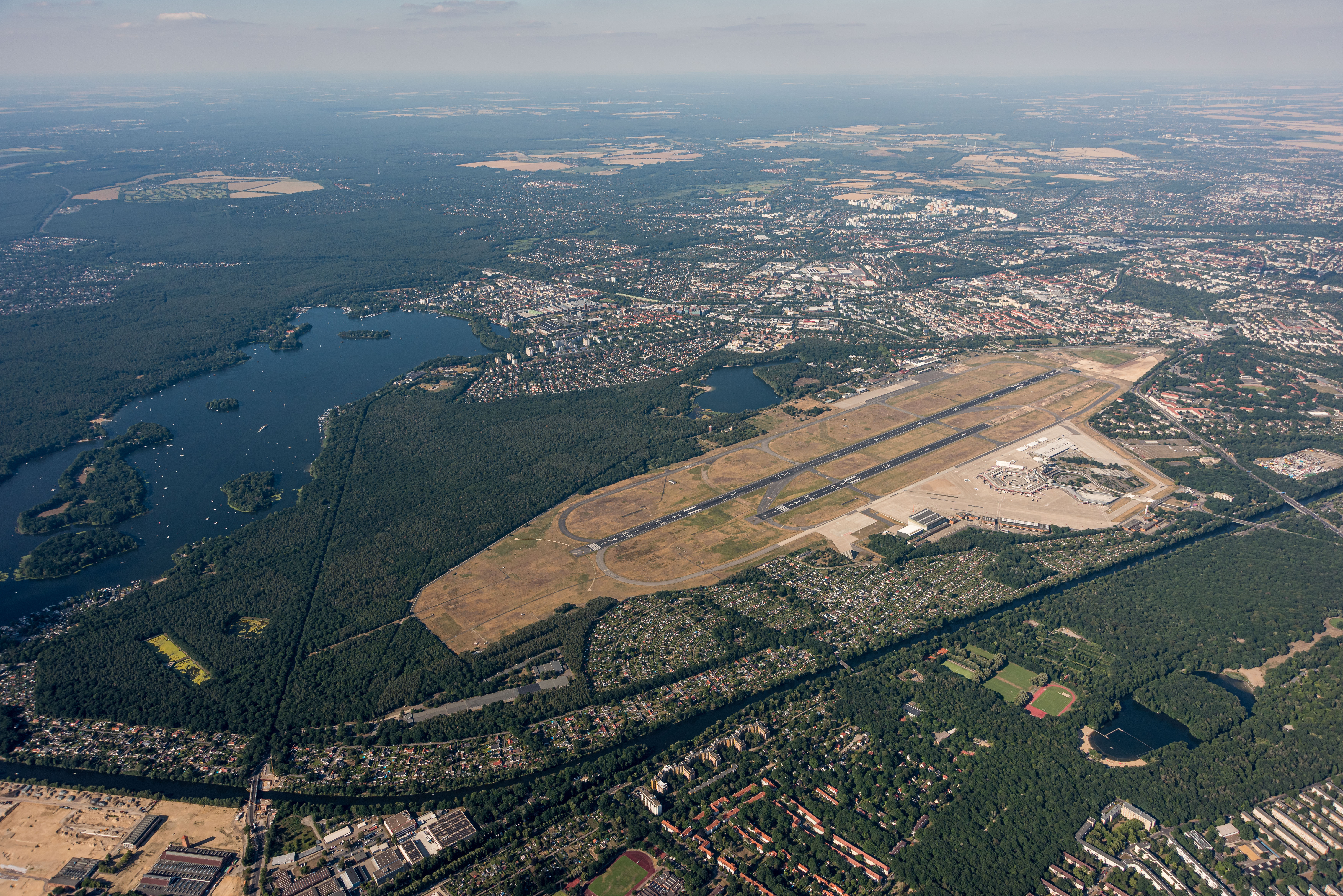 Luftaufnahme des ehemaligen Flughafens Berlin-Tegel mit Start- und Landebahn, Terminalgebäuden sowie den umliegenden Wäldern, Seen und Stadtgebieten.