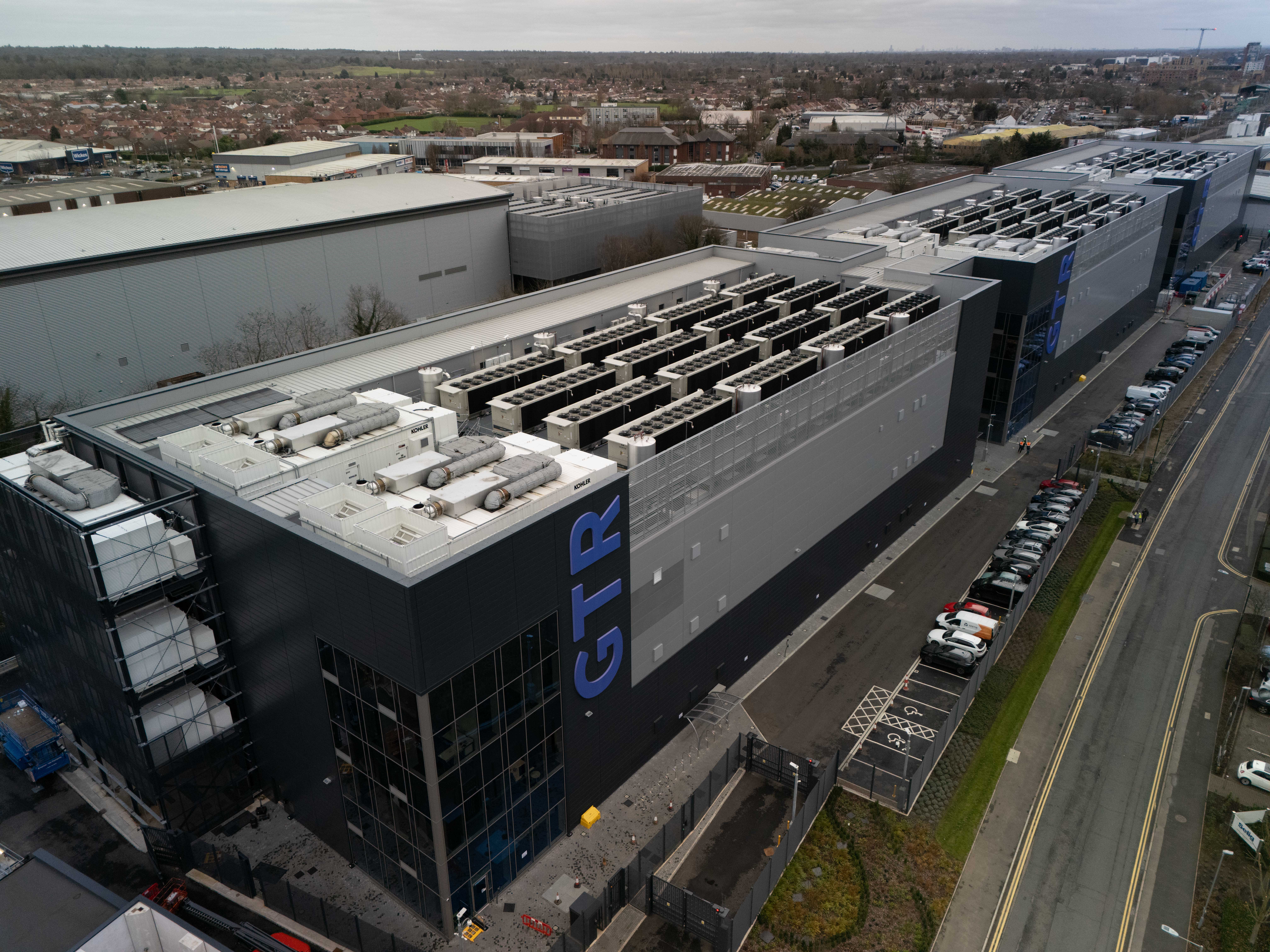 Oblique aerial view of the three GB One buildings with equipment on their roofs. A road and parking spaces run along the right.