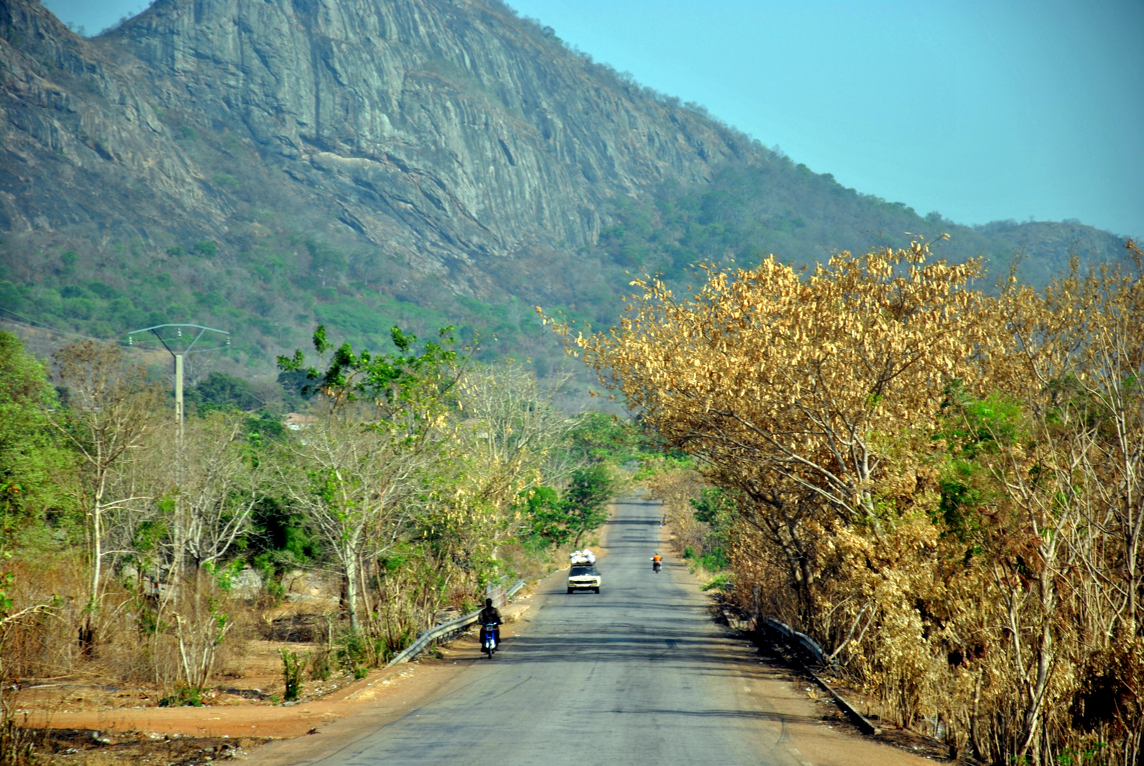 A concrete road between green yellowish leaves leading towards a green mountain in the back, rather far away on the long road a loaded car and two motorcycles 