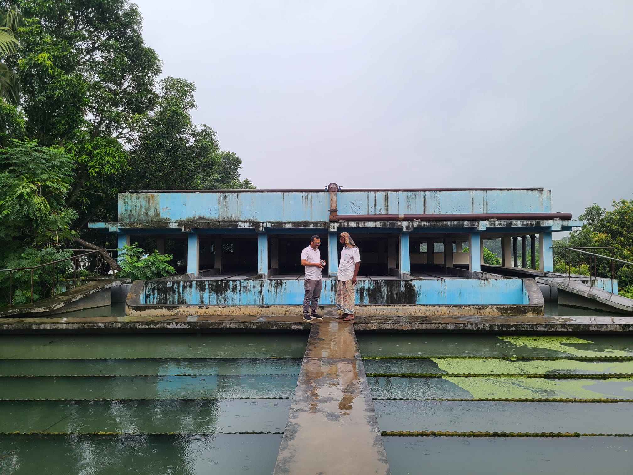 Two men facing each other, standing at in front of a blue building in the back of the picture, in front of them a well structured water basin.