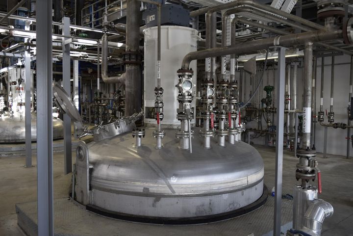 Large stainless-steel vessel with an open access hatch, valves and connected pipes inside a production hall.