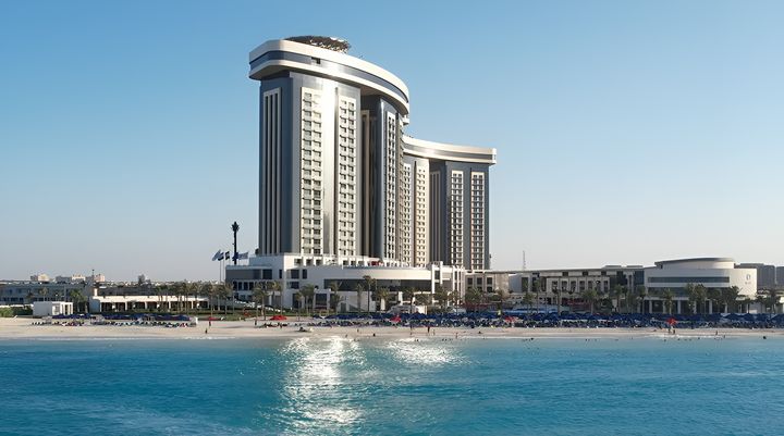 Blick vom Mittelmeer auf die Hoteltürme des Rixos Al Alamein mit vorgelagertem Sandstrand und Palmen.
