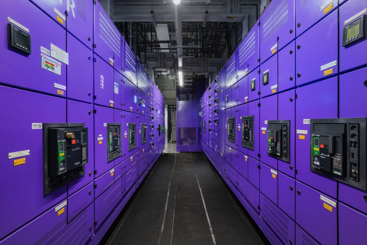 Aisle between two rows of purple electrical cabinets with black switches and control panels. Cables and lights run overhead.