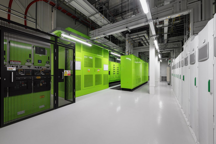 Interior plant room with large green equipment housings, a black safety enclosure on the left and a row of white cabinets on the right. Cable trays run overhead.